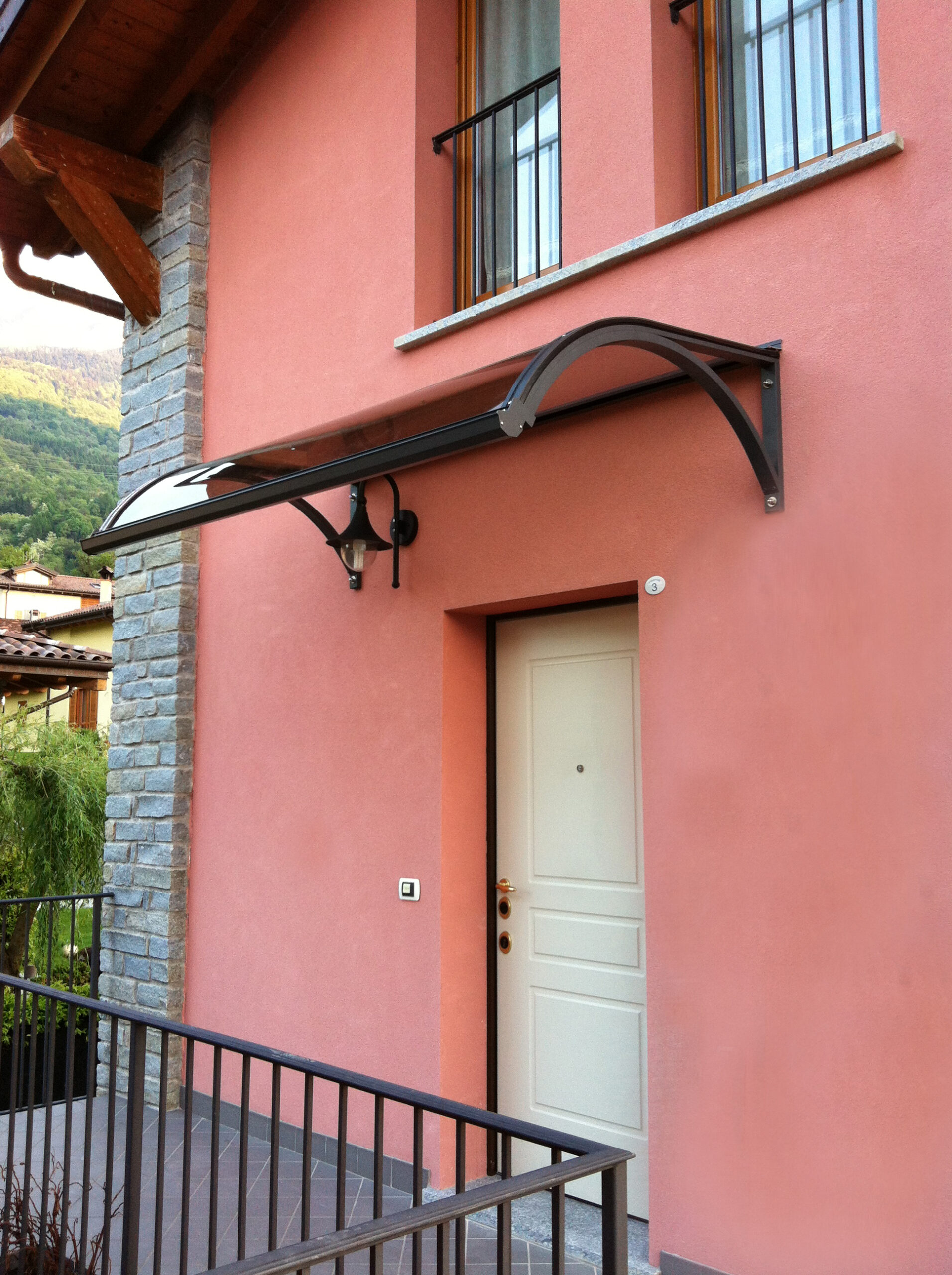 Pensilina curva P3 scura su ingresso in villaggio di montagna Pensilina curva P3 con struttura scura e lastra fumé installata sopra la porta d'ingresso di una casa in un villaggio di montagna