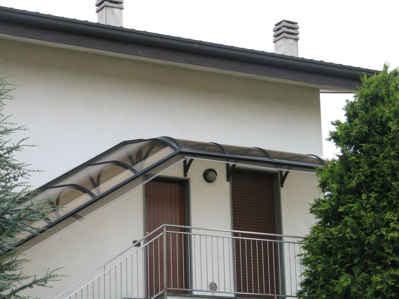 Pensilina curva P3 scura su terrazzo con cipressi Pensilina curva P3 con struttura scura e lastra fumé installata su un terrazzo con cipressi sullo sfondo
