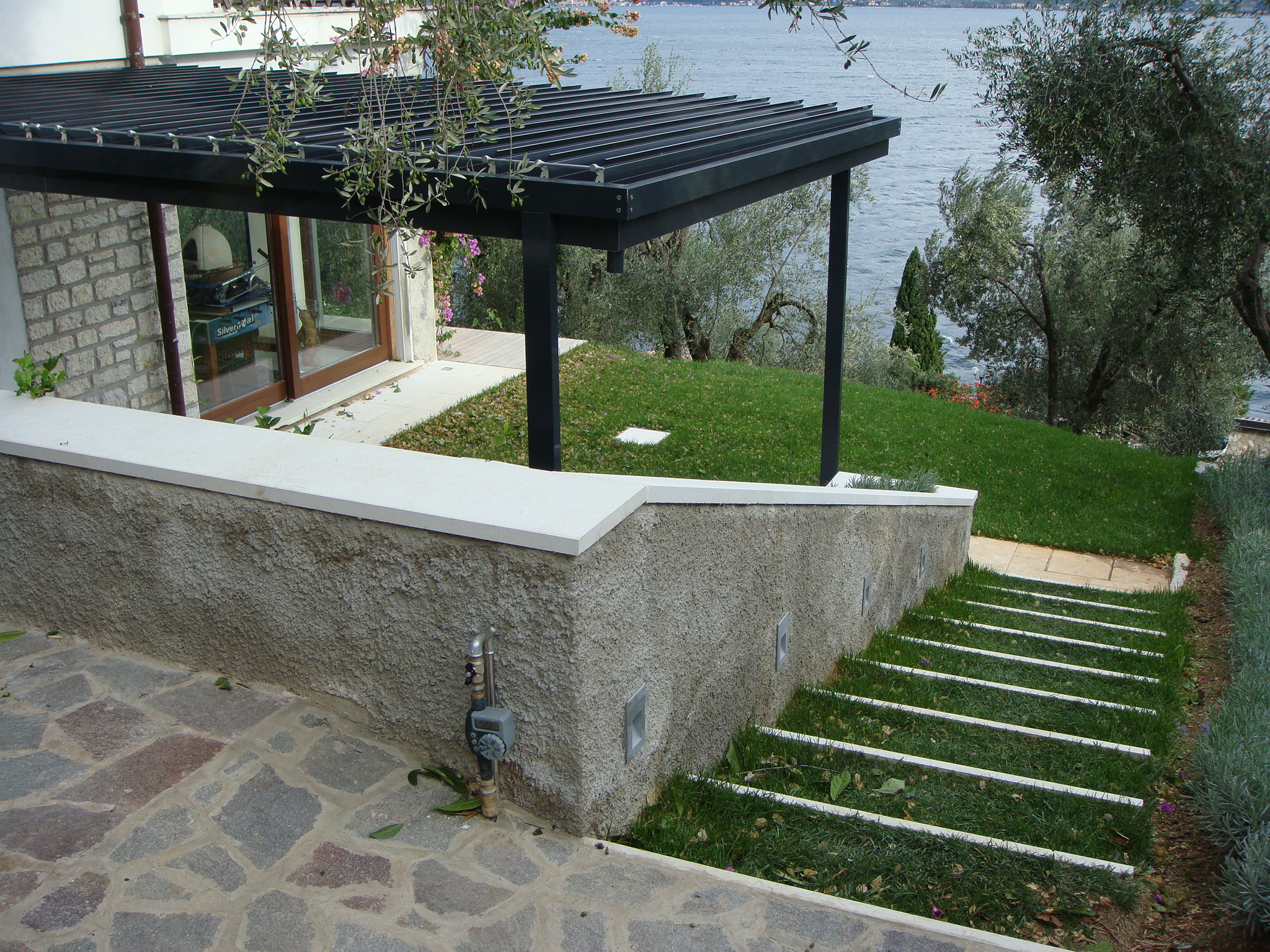 Pergola bioclimatica Ouverture scura affacciata sul lago Pergola bioclimatica Ouverture con struttura scura installata su una terrazza con vista sul lago e ulivi