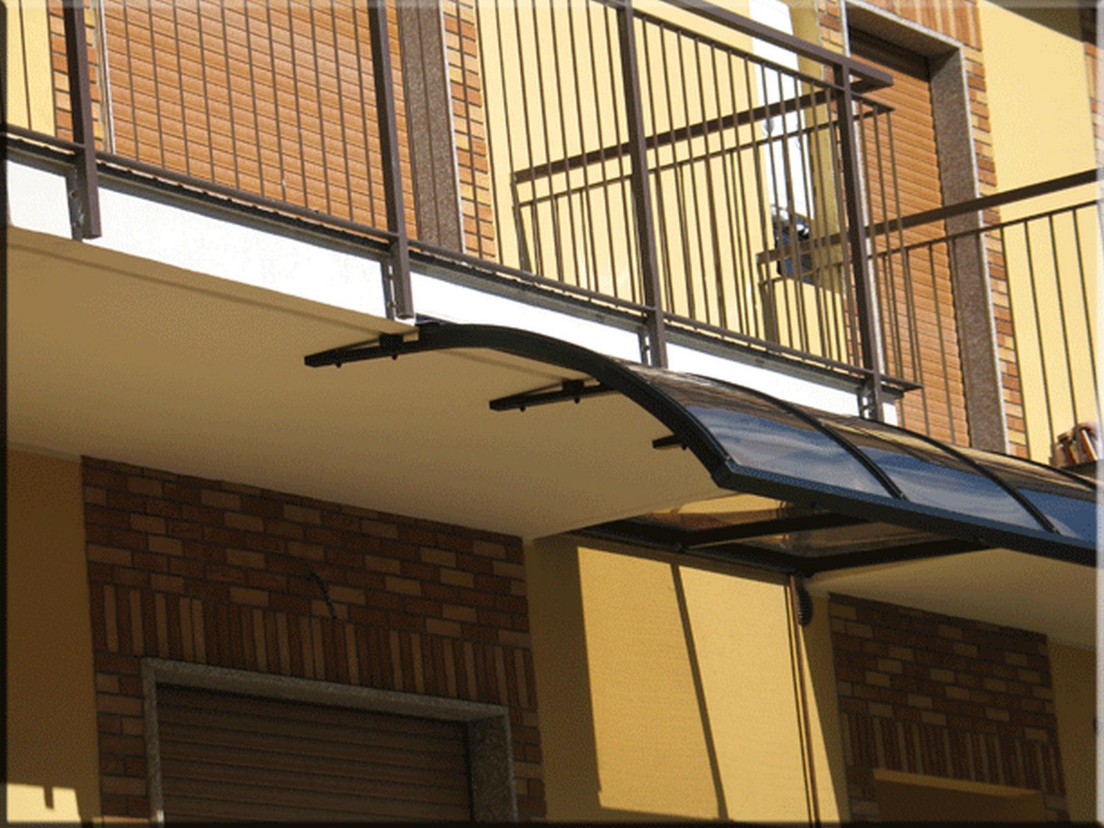 Pensilina sottobalcone scura installata su edificio residenziale Pensilina sottobalcone con struttura scura e lastra fumé installata sotto un balcone con ringhiera in ferro