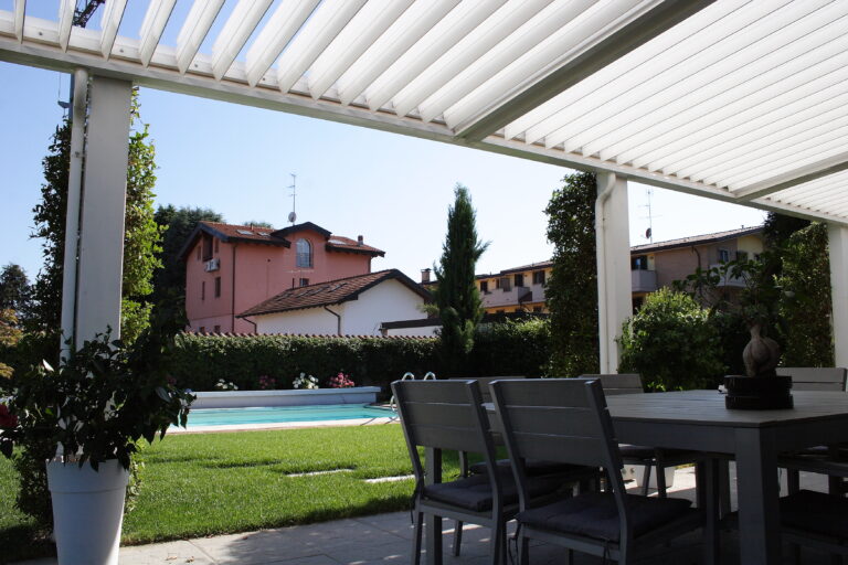 Pergola bioclimatica Ouverture bianca installata su una terrazza con vista su piscina e giardino